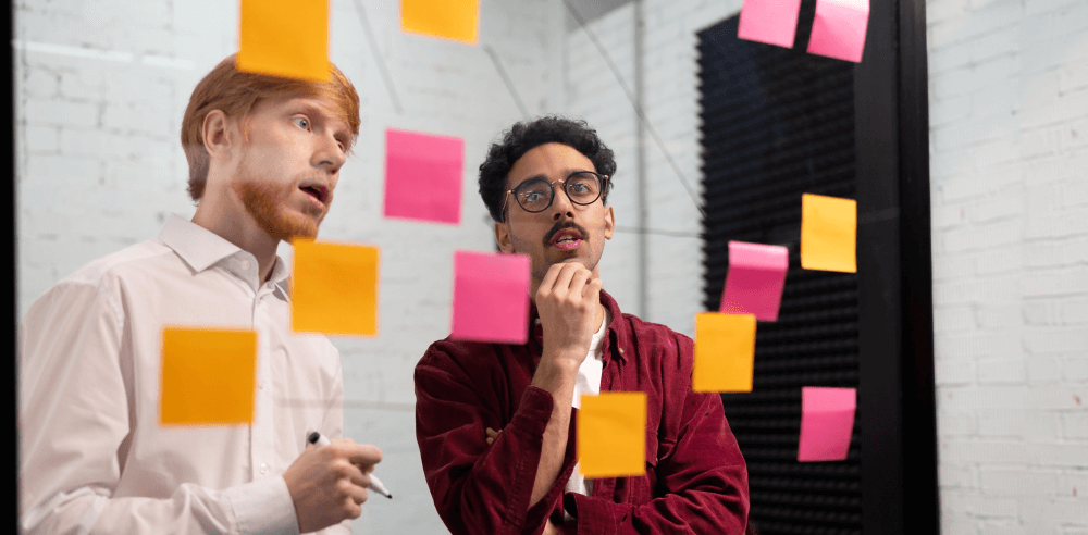 Two professionals brainstorming and discussing ideas in front of a whiteboard covered with colorful sticky notes.